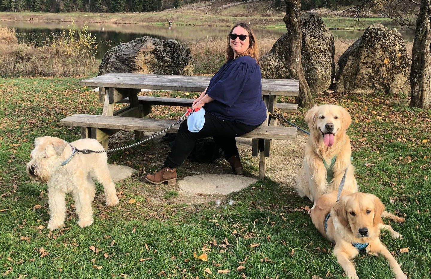 Woman with three dogs outside at a picnic bench on grass by a lake